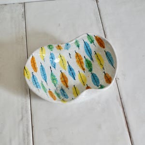 May include: A white, kidney-shaped ceramic dish with a colorful feather pattern. The feathers are in shades of blue, yellow, orange, and green. The dish has a slightly textured surface and is set against a white background.