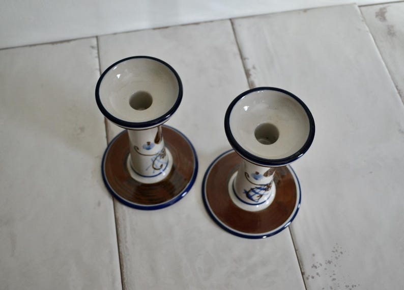 Puede incluir: Dos candelabros de cer&aacute;mica con un dise&ntilde;o floral marr&oacute;n, azul y blanco. Los candelabros tienen una base ancha y una parte superior estrecha con un agujero para una vela.