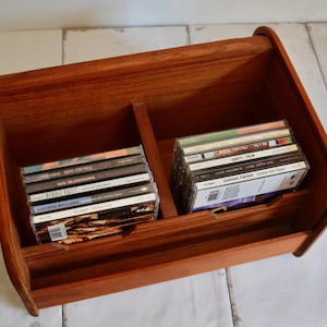May include: A medium-toned wooden storage rack with a curved top and a central divider. The rack features two open compartments, each containing a stack of compact discs with visible spine text including titles like Boys for Pele and Come On Over.