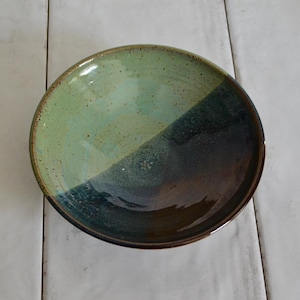 May include: A handmade ceramic bowl with a two-tone glaze. The upper half is a speckled light green, transitioning to a darker teal and brown on the lower half. The rim has a dark brown border. The bowl is round and sits on a white surface.