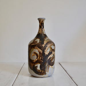 May include: A ceramic bottle-shaped vase with a narrow neck and rounded body. The vase features a light blue base color with brown and gold abstract designs. The vase is displayed on a white surface.