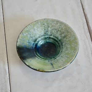 May include: A handmade ceramic bowl with a unique crystalline glaze. The bowl features a light green exterior with a dark blue interior. The glaze has a textured, almost frosted appearance, adding visual interest to the piece.