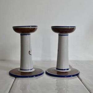 Puede incluir: Un par de candelabros de cer&aacute;mica blanca con detalles pintados en marr&oacute;n y azul. Los candelabros tienen una base ancha y una secci&oacute;n central estrecha.