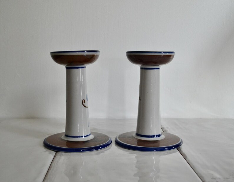 Puede incluir: Un par de candelabros de cer&aacute;mica blanca con detalles pintados en marr&oacute;n y azul. Los candelabros tienen una base ancha y una secci&oacute;n central estrecha.