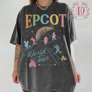 May include: Dark grey t-shirt with the word "EPCOT" in orange, green, pink, and yellow. The shirt features a graphic of the EPCOT sphere, cartoon characters, and the words "World Tour" and "Est. 1982".