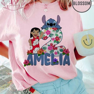 Op de afbeelding: Roze T-shirt met een afbeelding van Lilo en Stitch, het nummer 2, bloemmotieven en de naam "AMELIA". Het ontwerp bevat Stitch en Lilo in een rode jurk. Het shirt wordt gedragen met een blauwe short.