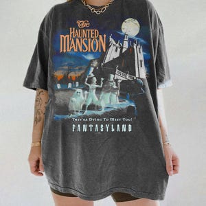 May include: Dark gray t-shirt featuring a graphic from The Haunted Mansion ride. The design includes the attraction's name, a spooky mansion, and ghostly figures. The text "They're Dying To Meet You!" and "FANTASYLAND" are also visible.