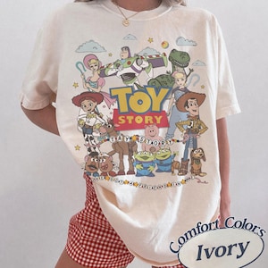 May include: An ivory short-sleeve t-shirt featuring a colorful graphic print of Toy Story characters including Woody, Buzz Lightyear, Jessie, and others. The design includes the Toy Story logo and the text I've Got A Friend In Me in a circular pattern.