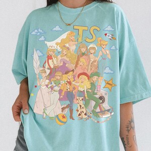May include: A light blue t-shirt featuring a graphic print of various animated characters in a collage style. The design includes the letters TS, a rocket, a sheriff star, a ball, and a camera. The characters are depicted with blonde hair.