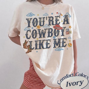 May include: An ivory t-shirt featuring the dark blue text YOU'RE A COWBOY LIKE ME. The graphic includes illustrations of brown cowboy boots, a red cowboy hat, a rocket, a yellow ball, and alphabet blocks with the letters T, S, and 5 among blue clouds.