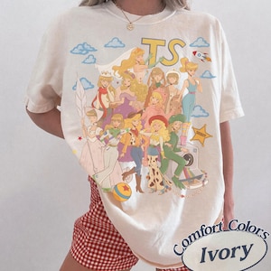 May include: An ivory short-sleeve t-shirt featuring a large graphic print of various animated characters in diverse outfits, including a space suit, cowboy attire, and gowns. The text TS is printed at the top, with clouds, a rocket, and a sheriff star.
