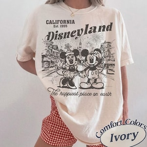 May include: Ivory-colored t-shirt featuring a vintage Disneyland graphic. The design includes Mickey and Minnie Mouse, fireworks, and the text "California Est. 1955." The phrase "The happiest place on earth" is also printed on the shirt.