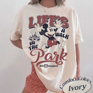 K&ouml;nnte beinhalten: Elfenbeinfarbenes T-Shirt mit einer Mickey Mouse-Grafik und dem Text "Life's a Walk in the Park" in Rot und Blau. Das Design beinhaltet ein Schloss und die Aufschrift "Walt Disney World". Das Shirt geh&ouml;rt zur Comfort Colors-Kollektion.
