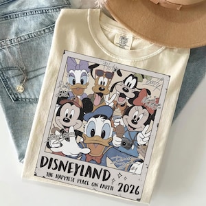 K&ouml;nnte beinhalten: Cremefarbenes T-Shirt mit einem Vintage-Disneyland-Grafikdesign mit Micky Maus, Minnie Maus, Donald Duck und anderen Disney-Figuren. Die Grafik enth&auml;lt den Text "Disneyland" und "The Happiest Place on Earth 2026."