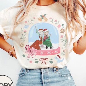 Puede incluir: Camiseta de color marfil con un diseño de globo de nieve con dos personajes de dibujos animados con gorros de Papá Noel. El globo de nieve está rodeado de ilustraciones navideñas, incluyendo campanas, flores y un bastón de caramelo. La camiseta es de material suave.