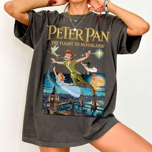 May include: Dark grey t-shirt with a colourful graphic from the film "Peter Pan." The graphic depicts Peter Pan, Wendy, Tinkerbell, and John flying over London. The text "Peter Pan The Flight to Neverland!" is printed in gold.