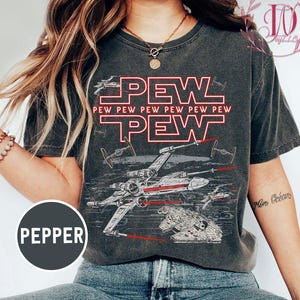 May include: Dark grey t-shirt with a graphic design featuring the word "PEW" in red, along with "PEW PEW PEW PEW PEW" and illustrations of spaceships. The shirt has a vintage, worn look.