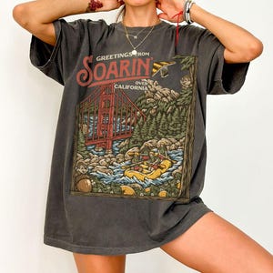 May include: Dark grey t-shirt with a graphic print of the Golden Gate Bridge, a river with people rafting, and the words "Greetings From Soarin' Over California". The design includes cartoon characters and a landscape.