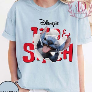 Op de afbeelding: Lichtblauw T-shirt met het Disney Stitch-personage. Het ontwerp toont Stitch die door een gescheurd papiereffect breekt met het woord "Stitch" in het rood. Het shirt is een casual kledingstuk met korte mouwen.