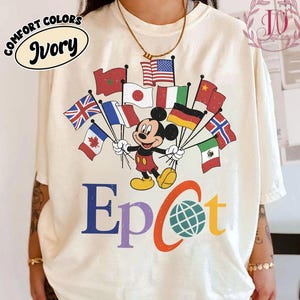 May include: Ivory-colored t-shirt featuring Mickey Mouse holding flags from various countries. The word "Epcot" is printed in blue, orange, and green. The shirt also has the words "Comfort Colors" and "Ivory" printed on it.