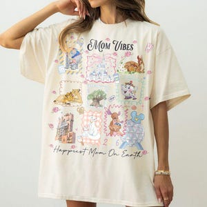 Puede incluir: Camiseta color crema con el texto "Mom Vibes" y "Happiest Mom On Earth". La camiseta presenta ilustraciones de dibujos animados coloridos de varios personajes y lugares emblemáticos. La camiseta tiene mangas cortas y un corte holgado.