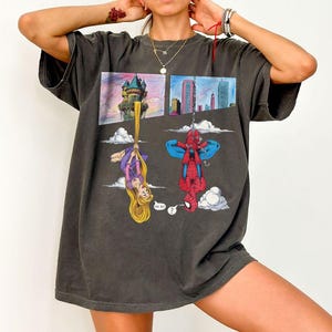 Peut inclure: T-shirt gris fonc&eacute; avec un motif color&eacute; de Raiponce et Spider-Man suspendus &agrave; l'envers. Raiponce est suspendue par ses longs cheveux blonds, et Spider-Man par une toile d'araign&eacute;e. Le t-shirt a un aspect vintage et us&eacute;.