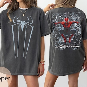 May include: Dark gray t-shirt featuring a large light blue spider graphic on the front and a Spider-Man graphic on the back. The back graphic includes the text "THE AMAZING SPIDER-MAN". The shirt is a relaxed fit.
