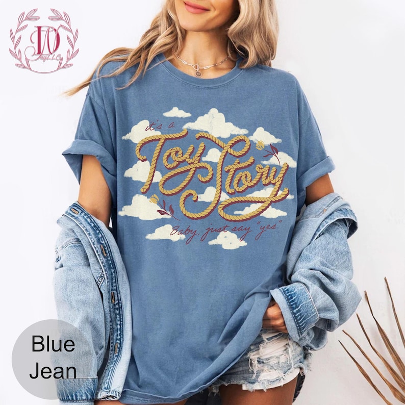 May include: A blue short-sleeve t-shirt featuring a graphic of white clouds and the text "It's a Toy Story" written in a yellow rope-style font, with the additional text "Baby, just say 'yes'" printed below in a smaller dark red script font.