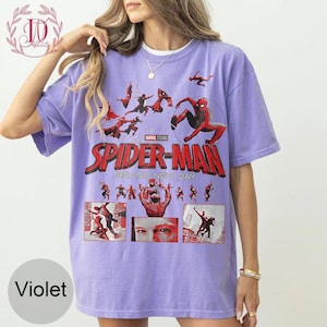 May include: Violet-colored t-shirt featuring the Spider-Man logo and various action poses of the superhero. The shirt includes the text "Marvel Studios" and "Grand New Day". The design is printed on a short-sleeved, crew neck tee.