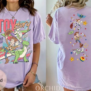 May include: A lilac t-shirt with a Toy Story design. The front has the Toy Story logo and characters Woody, Buzz, and Rex. The back features various characters and elements from the film, including rockets and stars. The text reads "TOY STORY" and "SINCE 1995".