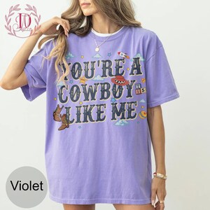 May include: A violet short-sleeved t-shirt featuring the text "YOU'RE A COWBOY LIKE ME" in a distressed dark blue font. The graphic includes illustrations of brown cowboy boots, a red cowboy hat, a rocket, stars, clouds, and a colorful ball.
