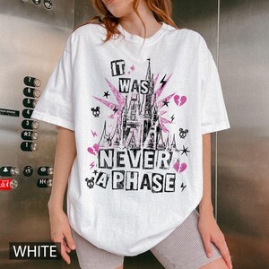 May include: White t-shirt with a graphic design featuring a pink castle, lightning bolts, stars, and the text "IT WAS NEVER A PHASE" in black and pink. The shirt has a crew neck and short sleeves.