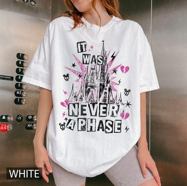 May include: White t-shirt with a graphic design featuring a pink castle, lightning bolts, stars, and the text "IT WAS NEVER A PHASE" in black and pink. The shirt has a crew neck and short sleeves.