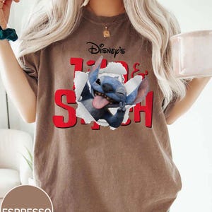 Op de afbeelding: Espresso-kleurig T-shirt met de tekst "Disney's Lilo & Stitch" in het rood. De afbeelding toont Stitch, een blauwe alien, die door een gescheurd papiereffect breekt. Een casual kledingstuk.