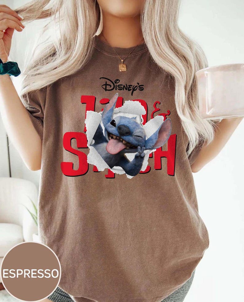 Op de afbeelding: Espresso-kleurig T-shirt met de tekst "Disney's Lilo & Stitch" in het rood. De afbeelding toont Stitch, een blauwe alien, die door een gescheurd papiereffect breekt. Een casual kledingstuk.