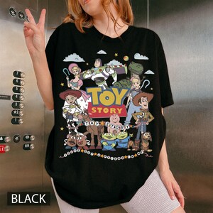 May include: A black t-shirt featuring a colorful graphic print of Toy Story characters including Woody, Buzz Lightyear, Jessie, and others. The design includes the text Toy Story and the phrase You've Got A Friend In Me, surrounded by clouds and stars.