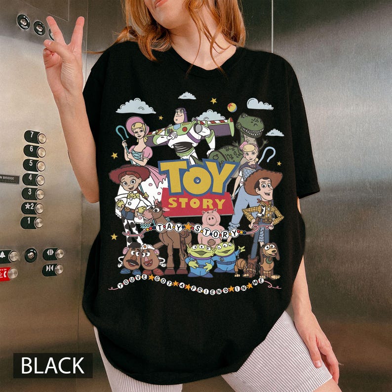 May include: A black t-shirt featuring a colorful graphic print of Toy Story characters including Woody, Buzz Lightyear, Jessie, and others. The design includes the text Toy Story and the phrase You've Got A Friend In Me, surrounded by clouds and stars.