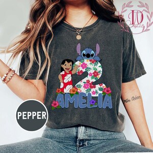 Op de afbeelding: Donkergrijs t-shirt met een kleurrijke afbeelding van Lilo en Stitch, het nummer 2, bloemmotieven en de naam "AMELIA" in blauwe letters. Het shirt heeft een vintage, versleten uitstraling.