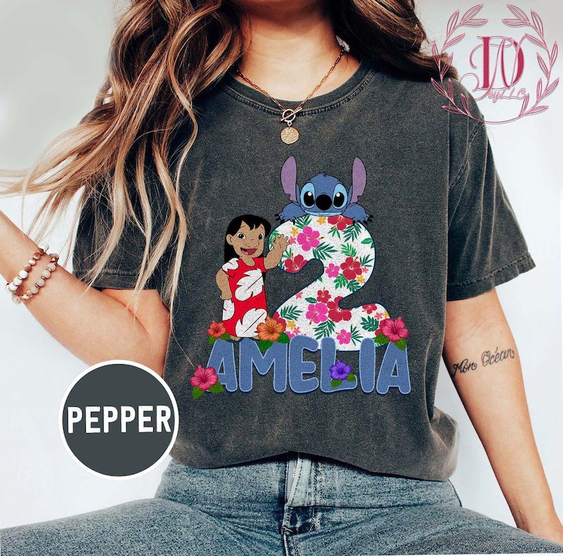 Op de afbeelding: Donkergrijs t-shirt met een kleurrijke afbeelding van Lilo en Stitch, het nummer 2, bloemmotieven en de naam "AMELIA" in blauwe letters. Het shirt heeft een vintage, versleten uitstraling.