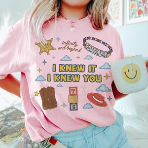 Puede incluir: Una camiseta rosa claro con el texto I KNEW IT I KNEW YOU en amarillo y azul. Los gr&aacute;ficos incluyen una estrella de sheriff con TS, el texto infinity and beyond, una pulsera de cuentas con YOU'VE GOT A FRIEND IN ME, botas, bloques y un sombrero.