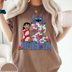 Op de afbeelding: Espresso-kleurig T-shirt met een kleurrijke afbeelding van een tekenfilmfiguur, een meisje en het cijfer 2 versierd met bloemen. De naam "AMELIA" staat eronder in blauwe letters.