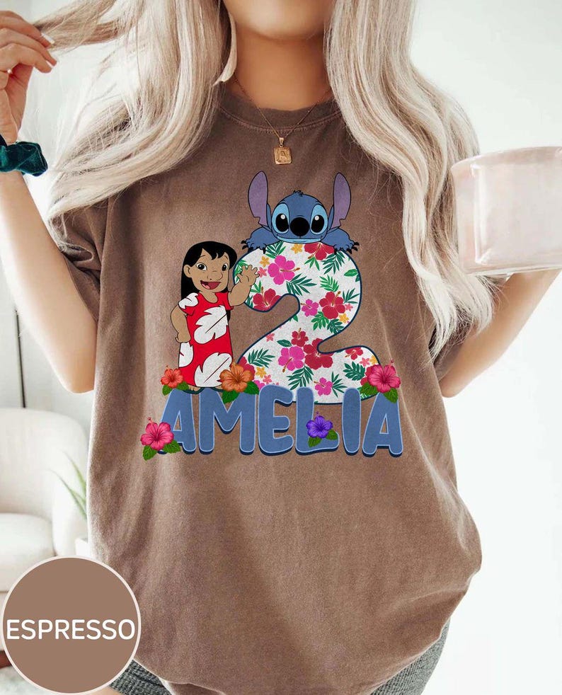 Op de afbeelding: Espresso-kleurig T-shirt met een kleurrijke afbeelding van een tekenfilmfiguur, een meisje en het cijfer 2 versierd met bloemen. De naam "AMELIA" staat eronder in blauwe letters.