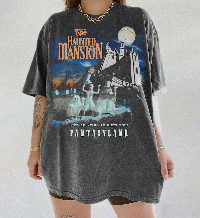 May include: Dark gray t-shirt featuring a graphic from The Haunted Mansion ride. The design includes the attraction's name, a spooky mansion, and ghostly figures. The text "They're Dying To Meet You!" and "FANTASYLAND" are also visible.