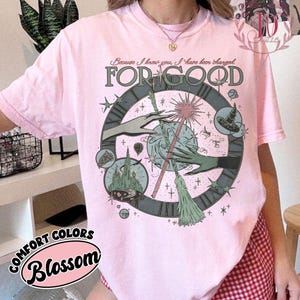 Puede incluir: Una camiseta rosa claro con un dise&ntilde;o gr&aacute;fico que presenta una esfera de reloj, manos y una varita m&aacute;gica. El texto "FOR GOOD" se muestra de forma destacada, con texto adicional arriba. La camiseta est&aacute; etiquetada como "COMFORT COLORS Blossom".
