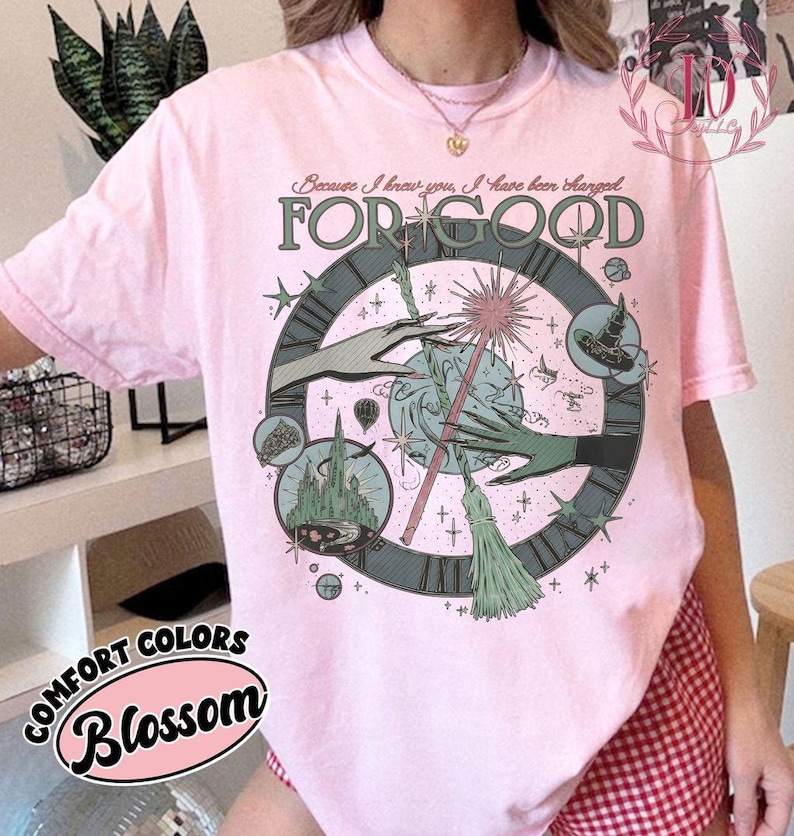 Puede incluir: Una camiseta rosa claro con un dise&ntilde;o gr&aacute;fico que presenta una esfera de reloj, manos y una varita m&aacute;gica. El texto "FOR GOOD" se muestra de forma destacada, con texto adicional arriba. La camiseta est&aacute; etiquetada como "COMFORT COLORS Blossom".