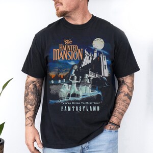 May include: Black t-shirt featuring a graphic from "The Haunted Mansion" ride at Disneyland's Fantasyland. The design includes the ride's logo, a spooky mansion, and ghostly figures under a full moon. Text reads "They're Dying To Meet You!"