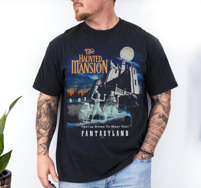 May include: Black t-shirt featuring a graphic from "The Haunted Mansion" ride at Disneyland's Fantasyland. The design includes the ride's logo, a spooky mansion, and ghostly figures under a full moon. Text reads "They're Dying To Meet You!"