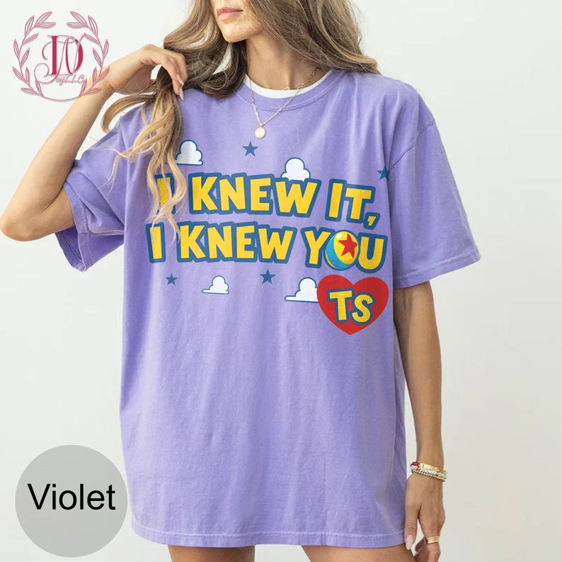 May include: A violet short-sleeve t-shirt featuring the yellow text "I KNEW IT, I KNEW YOU" with a colourful ball graphic replacing the letter O, a red heart containing the letters "TS", and white cloud and blue star accents printed on the front.