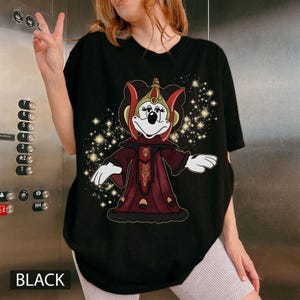 Puede incluir: Camiseta negra con un personaje de dibujos animados con un atuendo rojo y dorado y un fondo estrellado. El personaje tiene una cara blanca y lleva una corona. La palabra "BLACK" est&aacute; impresa en la parte inferior de la camiseta.