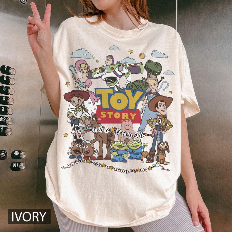 May include: An ivory t-shirt featuring a colorful graphic print of Toy Story characters including Woody, Buzz Lightyear, Jessie, and others. The central design includes the Toy Story logo and the text You've Got A Friend In Me in a decorative font.